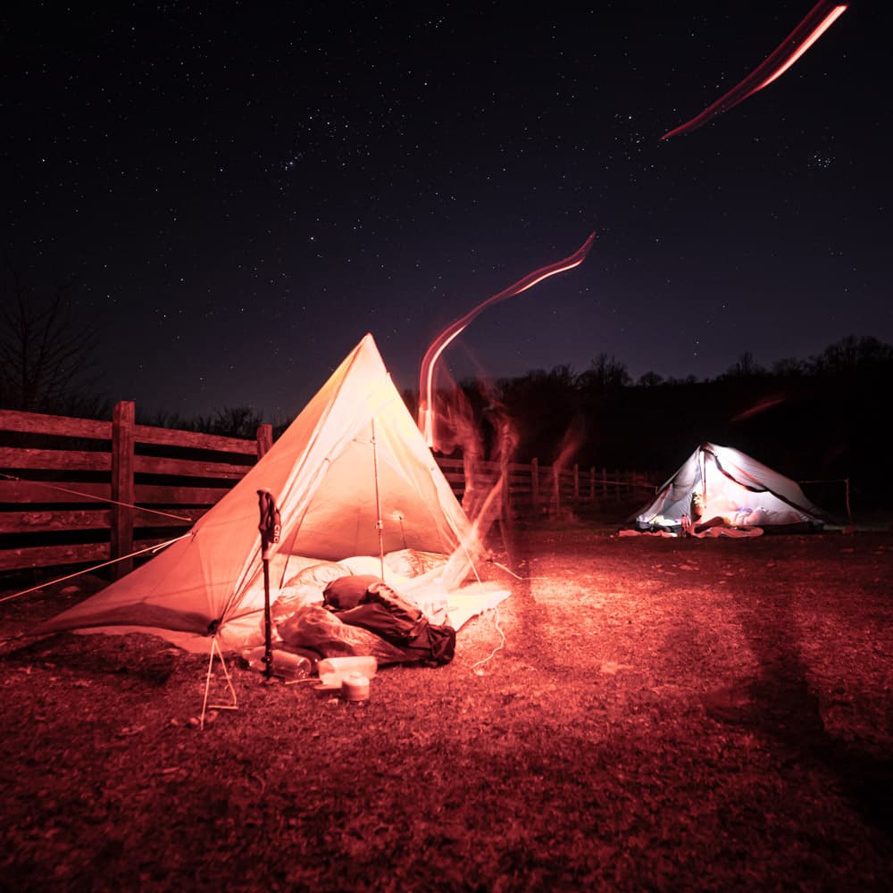 2 tents lit by red and white headlamps on a starry night setup next to a horse fence