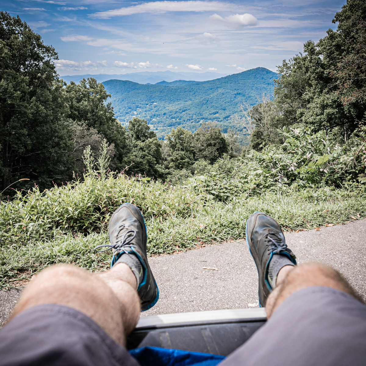 First person view of a hikers legs and feet extended towards the middleground which are a tree-cropped view of the Appalacian mountains