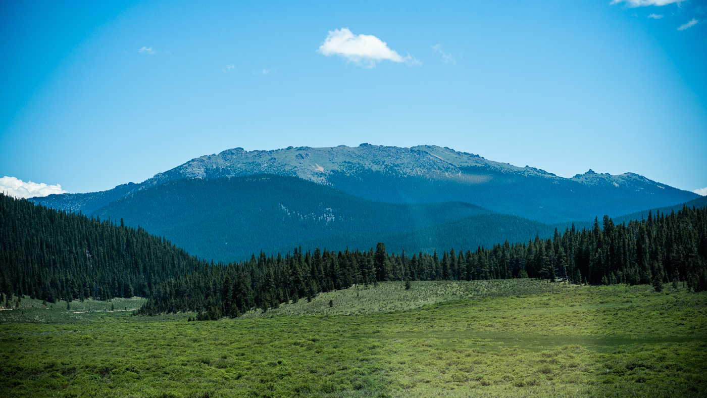 A vast Colorado mountain landscape spreads the view with 4 layers: sky at the top, hazy opaque round mountains, pointy evergreen trees, meadow at the bottom