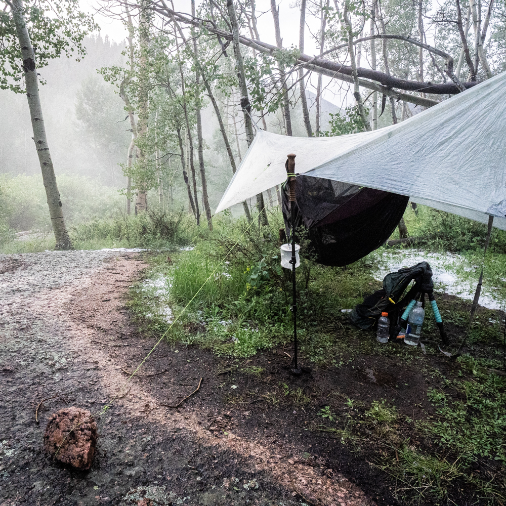 A hammock + tarp shelter hangs in a misty damp landscape, around the tarp edges hale lies