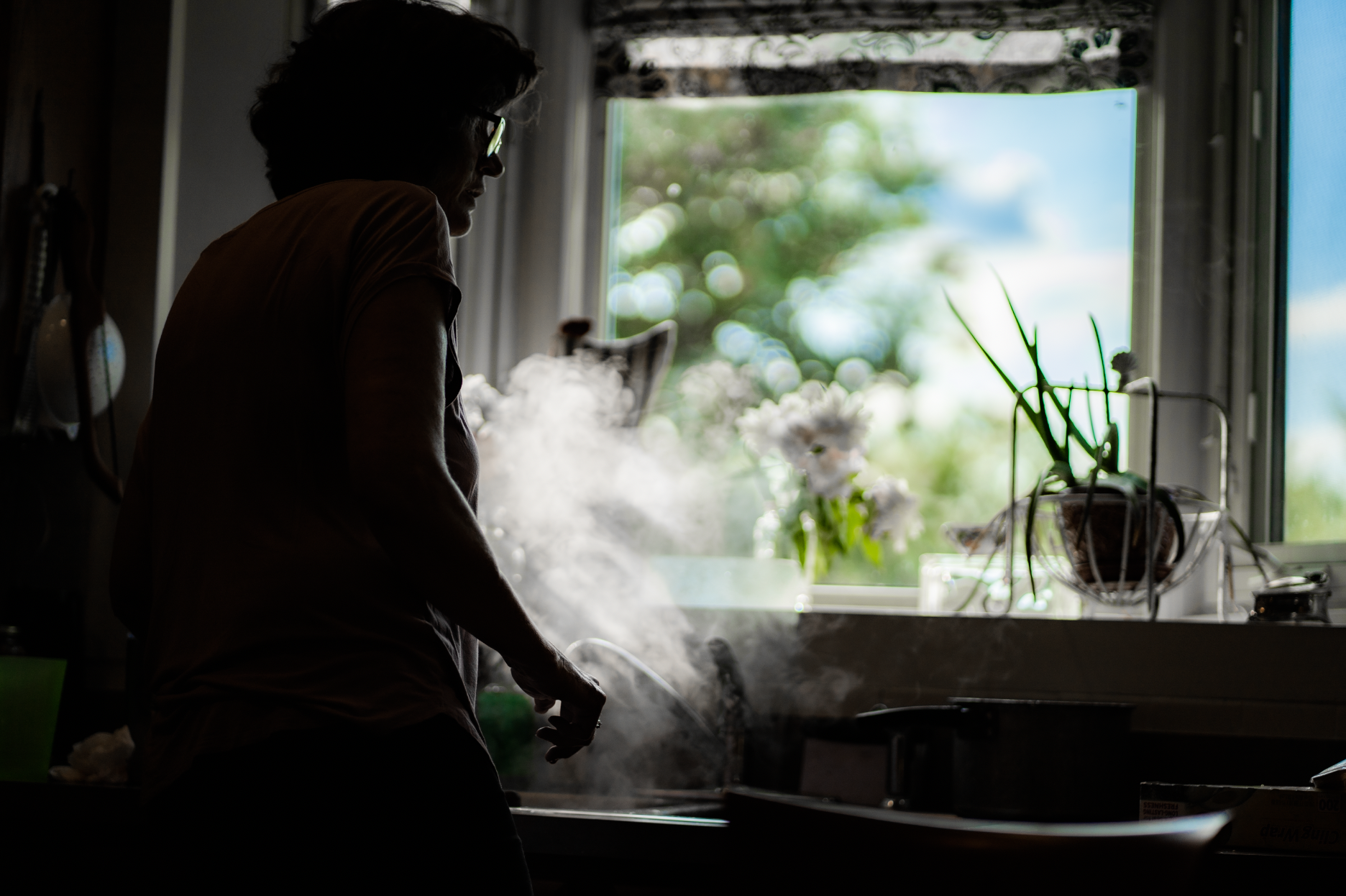 Silhouette of a hurried woman looking away from a kitchen sink that is steaming from cooked pasta draining, afternoon light flooding in from a window behind