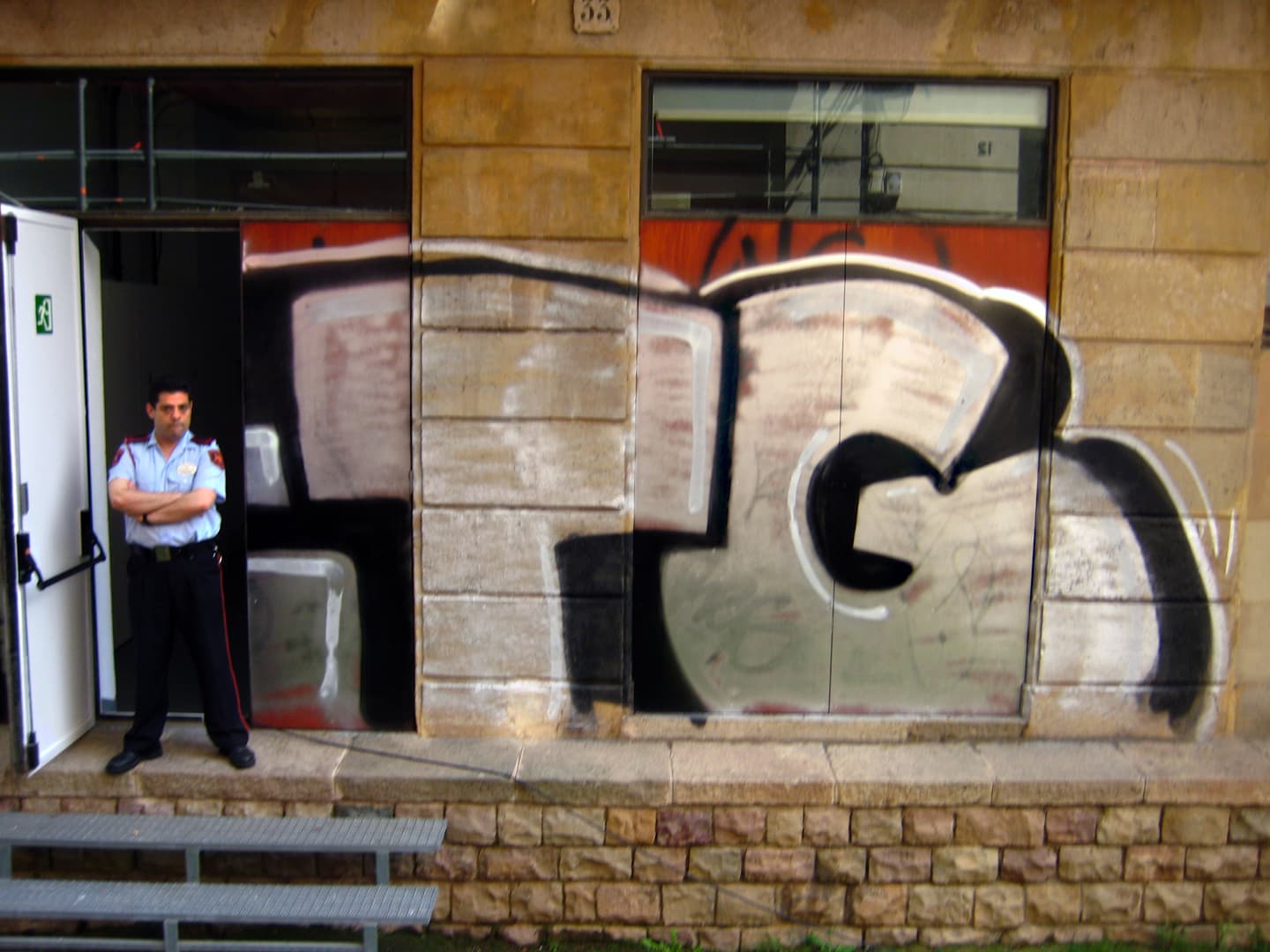 Security guard outside an open door to a large building with his arms crossed and with graffitti blocked out on the wall behind him