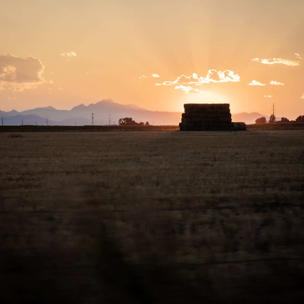 Beams of bright light silhouette a barn in the foreground, background is the Rocky mountains drenched in golden sunset light